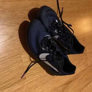 Svarta Nike löparskor med spik - Säljer ett par svarta Nike löparskor med spik, perfekta för friidrott. Skorna har en vit swoosh och lila detaljer på sulan. De är i mycket bra skick och kommer med en praktisk förvaringspåse. Perfekta för träning på bana!