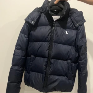 Marinblå pufferjacka från Calvin Klein - Säljer en snygg mörkblå pufferjacka från Calvin Klein i ett sprillans nytt skick. Jackan har en dragkedja framtill och är quiltad för extra värme. Den har långa ärmar och en huva för att skydda mot kyla. Perfekt för vintern och hösten!