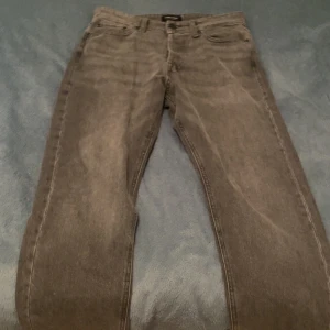 Grå jeans från Jack & Jones - Säljer ett par grå jeans från Jack & Jones i modellen Relaxed/Chris. De har en avslappnad passform och är perfekta för en casual look. Jeansen är i bra skick och har en klassisk design med knappar och dragkedja. Perfekta för både vardag och fest!