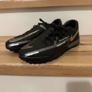 Nike Phantom fotbollsskor - Säljer ett par Nike Phantom fotbollsskor i svart och guld. Skorna har en snygg design med snörning och är perfekta för spel på konstgräs. De är i mycket bra skick och redo för nästa match!