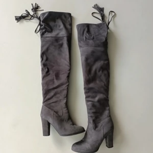 Grå mockastövlar med klack - Snygga grå stövlar i mocka med hög klack och dragkedja på sidan. De har en elegant spetsig tå och knytning upptill för extra stil. Perfekta för höst och vinter, både till vardags och fest.