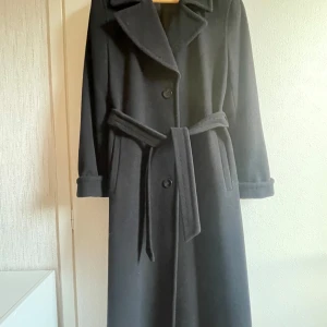 Svart kappa från Steilmann (Tyskland) - Säljer en elegant svart kappa från Steilmann i mycket bra skick. Den har långa ärmar och ett bälte i midjan för en snygg passform. Perfekt för höst och vinter, med klassiska knappar framtill. En tidlös stil som passar till alla tillfällen.