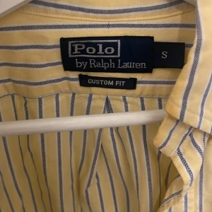 Gul och blå randig skjorta från Polo Ralph Lauren - Säljer en snygg gul och blå randig skjorta från Polo Ralph Lauren i storlek S. Skjortan har långa ärmar och en klassisk krage. Den är i bomull och har en broderad logga på bröstet. Perfekt för både vardag och fest! 💙💛