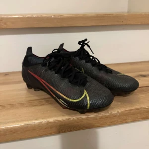 Nike mecurial elite fotbollskor svart - Säljer ett par svarta Nike fotbollsskor i bra skick. De har en snygg design med gula och röda detaljer och är perfekta för fotbollsträning. Skorna har snörning och en slitstark sula för bra grepp på planen.