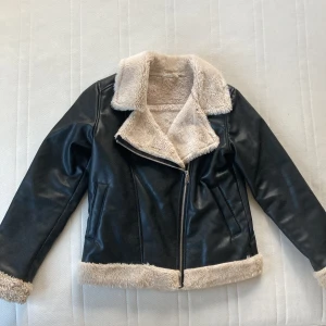 Svart skinnjacka med foder från H&M - Säljer en snygg svart skinnjacka från H&M med mjukt beige foder. Jackan har en dragkedja framtill och är perfekt för höst och vår. Den har långa ärmar och en cool stil som passar både till vardags och fest. Perfekt för att hålla sig varm och stilren! 🖤