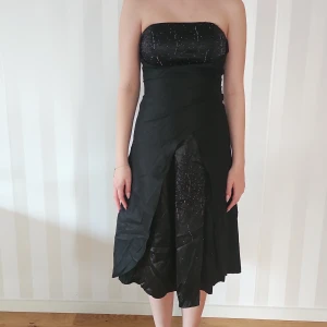 Svart axelbandslös klänning - Snygg svart axelbandslös klänning med glittriga detaljer. Perfekt för festliga tillfällen! Den har en elegant snörning i ryggen och en fin passform som framhäver figuren. Klänningen är knälång och har en stilren design.