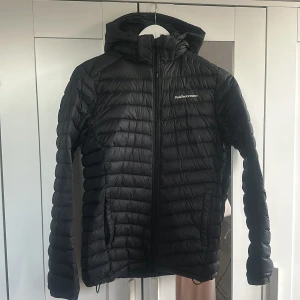 Svart dunjacka från Peak Performance - Säljer en svart dunjacka från Peak Performance i mycket bra skick. Jackan har en snygg design med dragkedja och huva, perfekt för kalla dagar. Den är lätt och bekväm att bära, idealisk för både vardag och utomhusaktiviteter. Säljer för 600kr.