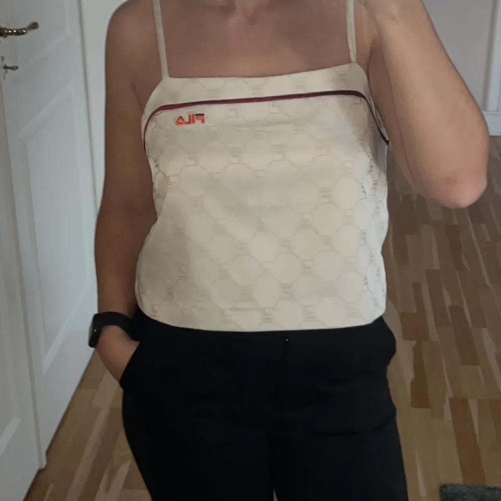 Säljer en snygg beige topp från Fila med ett diskret mönster och röd logga. Toppen har tunna axelband och en stilren design, perfekt för sommardagar eller en avslappnad fest. Materialet känns mjukt och bekvämt mot huden då tröja är fodrad. . Toppar.