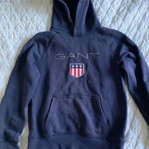 Mörkblå hoodie från GANT 146-152 cm - Säljer en snygg mörkblå hoodie från GANT med broderad logga och emblem på bröstet. Den har en klassisk känguruficka och är perfekt för både höst och vinter. Hoodien är i bra skick och superbekväm att ha på sig. Perfekt för en casual look! 💙