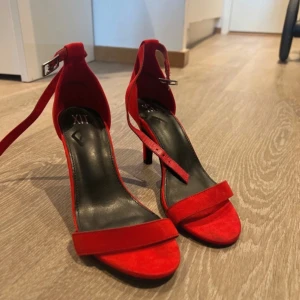 Röda klackskor med ankelrem - Snygga röda klackskor i mocka med en elegant ankelrem och öppen tå. Perfekta för festliga tillfällen eller en utekväll. De har en smal klack som ger en stilren look. Skorna är i mycket bra skick och redo att användas för att lyfta din outfit. Endast använd 1 gång.