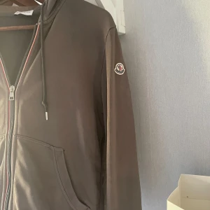 Brun hoodie med dragkedja från Moncler - Säljer en snygg brun hoodie från Moncler i mycket bra skick. Den har en dragkedja framtill och en praktisk huva med dragsko. Perfekt för höst och vår, med långa ärmar och en bekväm passform. Moncler-loggan pryder ärmen. Passar både till vardags och lite mer avslappnade tillfällen. Står att det är storlek medium men sitter som en xs. Har en liten skada vi fickan. Denna produkt är ej prutbart.