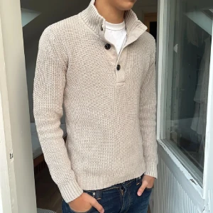 Beige stickad tröja med knappar - Säljer en snygg beige stickad tröja med långa ärmar och ribbad krage. Tröjan har två knappar vid halsen för en stilren look. Perfekt för höst och vinter, och passar både till vardags och lite finare tillfällen. Supermysig och varm!
