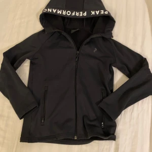Svart jacka från Peak Performance - Säljer en svart jacka från Peak Performance i mycket bra skick. Den har en dragkedja framtill och en huva med textdetaljer. Perfekt för höst och vår, med en stilren look och bekväm passform. Passar både till vardags och lättare träning. Kostar ca 1000kr ny