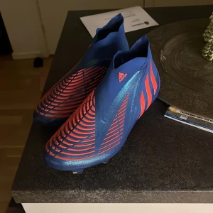 Blå och röda fotbollsskor från Adidas - Säljer ett par coola fotbollsskor från Adidas i blått och rött. Skorna har en modern design med mönstrade detaljer och är perfekta för fotbollsträning eller match. De är i mycket bra skick och har en tight passform för optimal kontroll på planen.