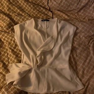 Beige blus från Zara - Säljer en elegant beige blus från Zara i storlek S. Blusen har en omlott-design med en knytning i midjan som ger en fin siluett. Perfekt för både vardag och festliga tillfällen. Materialet känns mjukt och bekvämt mot huden.
