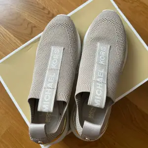 Snygga beige sneakers från Michael Kors i stickat material. De har en vit sula och guldiga detaljer vid hälen. Perfekta för en stilren look och superbekväma att ha på sig. Passar både till vardags och lite finare tillfällen. Märkesloggan syns tydligt på ovansidan.