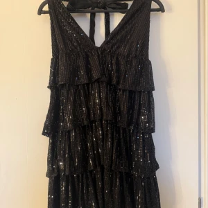 Svart glittrig klänning från Shein - Säljer en superfin svart klänning från Shein i storlek L. Den har ett glittrigt mönster och volanger som ger en festlig look. Perfekt för nyår eller en annan festlig tillställning. Klänningen är ärmlös och har en v-ringad design med knytning i nacken.