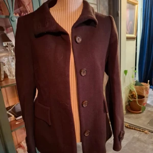 Brun kappa från Burberry - Säljer en elegant brun kappa från Burberry i storlek 42. Den har en klassisk design med knappar framtill och ett bälte bak för en snygg passform. Fodret har det ikoniska Burberry-rutiga mönstret. Perfekt för höst och vinter!