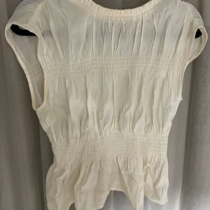 Creme vit blus med resårdetaljer - Säljer en söt vit blus med resårdetaljer i midjan och vid ärmslut. Blusen är ärmlös och har en luftig passform, perfekt för varma sommardagar. Materialet är lätt och bekvämt, vilket gör den idealisk för både vardag och festliga tillfällen.