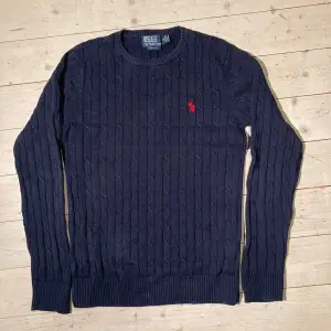 Säljer en snygg mörkblå stickad tröja från Polo Ralph Lauren. Den har ett klassiskt flätmönster och den ikoniska röda loggan på bröstet. Perfekt för både höst och vinter, med långa ärmar och ribbade kanter. Passar till både vardag och fest!