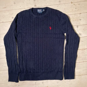 Mörkblå stickad tröja från Polo Ralph Lauren - Säljer en snygg mörkblå stickad tröja från Polo Ralph Lauren. Den har ett klassiskt flätmönster och den ikoniska röda loggan på bröstet. Perfekt för både höst och vinter, med långa ärmar och ribbade kanter. Passar till både vardag och fest!