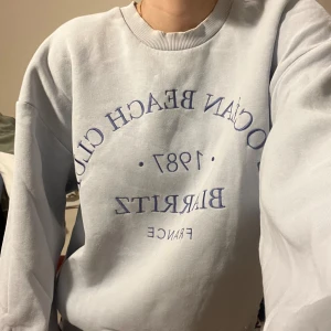 Ljusblå sweatshirt med tryck - Säljer en ljusblå sweatshirt med texten 'L'OCEAN BEACH CLUB 1987 BIARRITZ FRANCE' i mörkblått. Tröjan har en avslappnad passform och är perfekt för en chill dag. Den är långärmad och gjord i mjukt material. Perfekt för höst och vår!