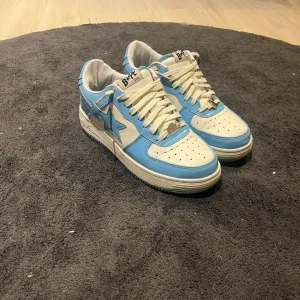Blå och vita sneakers från A Bathing Ape - Säljer ett par snygga sneakers från A Bathing Ape i blått och vitt. Skorna har en cool stjärndetalj på sidan och klassisk snörning. De är i mycket bra skick och kommer med originalkartong och påse. Perfekta för att ge din outfit en unik touch!