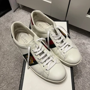 Vita sneakers från Gucci - Säljer ett par vita Gucci Ace sneakers med klassiska gröna och röda ränder samt ett broderat emblem på sidan. Skorna är i bra skick med helt nya skosnören och har lite slitage på innersulan. Perfekta för både vardag och fest!