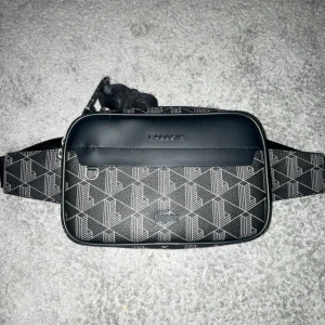 Svart midjeväska från Lacoste - Säljer en snygg svart midjeväska från Lacoste med geometriskt mönster. Väskan har en praktisk dragkedja och justerbart band. Insidan är fodrad med en cool orange detalj och har flera fack för enkel organisering. Perfekt för både vardag och fest!