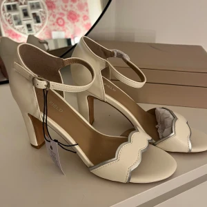 Krämvita klackskor med remmar - Säljer ett par fina krämvita klackskor som aldrig är använda. De har en söt vågig kantdetalj och justerbara remmar runt ankeln. Perfekta för fest eller bal! Skorna är storlek 37.