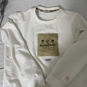 Vit långärmad tröja från Burberry - Säljer en stilren vit långärmad tröja från Burberry. Den har en snygg guldfärgad patch med Burberry-loggan på framsidan och klassiskt rutigt mönster på insidan av kragen. Perfekt för både vardag och fest! 🕶️