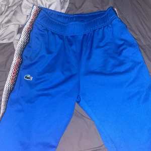 Blå träningsset från Lacoste - Säljer ett blått träningsset från Lacoste med vita och röda detaljer. Setet består av en jacka och byxor, båda med det klassiska Lacoste-märket. Perfekt för sport och träning.