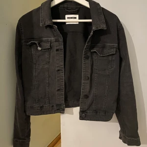 Svart jeansjacka  - Snygg svart jeansjacka med klassisk krage och knappar framtill. Jackan har två bröstfickor med lock och knapp samt långa ärmar. Perfekt för en cool och avslappnad stil. Skulle säga att den sitter mer som en S än en M