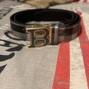 Rutig bälte med guldspänne. Burberry  - Snyggt bälte med ett stort B-format guldspänne. Bältet har ett stilrent rutigt mönster i svart, grått och vitt. Perfekt för att ge en touch av elegans till din outfit. Pris kan diskuteras vid snabbt köp