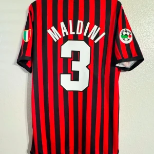 AC Milan Maldini XXL - Säljer en klassisk randig fotbollströja från Adidas med Maldini och nummer 3 på ryggen. Tröjan har korta ärmar och är i rött och svart med sponsorlogga från Opel på framsidan. Perfekt för samlare eller fans av italiensk fotboll.