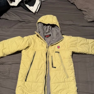 Gul vintage dunjacka från Fjällräven - Säljer en gul vintage dunjacka från Fjällräven med huva och dragkedja. Jackan har flera praktiska fickor med dragkedjor och är perfekt för kyligare väder. Den har en quiltad design och är långärmad. Den är storlek S och har lite små defekter så som lite smuts längst ner på den vänstra armen (se bild 4)