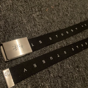 Svart bälte från Stussy - Snyggt svart bälte från Stussy med silverfärgat spänne. Bältet har Stussy-loggan tryckt längs hela remmen och på spännet. Perfekt för att ge en streetwear-look till din outfit. Bältet är 120cm långt
