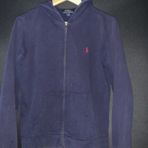 Mörkblå hoodie från Ralph Lauren - Säljer en snygg mörkblå hoodie från Ralph Lauren med dragkedja och det klassiska röda logotypen på bröstet. Strlk L passar till 152-168 men jag har vuxit ur den. Och JA tröjan är äkta jag har haft den i 1,5 år. Har ett litet hål i högra ärm. Syns inte om man inte kollar riktigt noga.