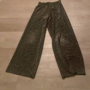 Glittriga byxor från KappAhl - Snygga glittriga byxor från KappAhl i en mörk färg. Byxorna har en wide passform och är perfekta för att sticka ut på festen. De är tillverkade i ett glittrigt material som ger en extra touch av glamour.