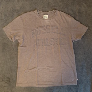 Russell Athletic tshirt  - Brun t-shirt från Russell Athletic med stort tryck på framsidan. Kortärmad och har en klassisk rund halsringning. Perfekt för att träna i då materialet är bomullsmix eller för att chilla hemma. Skriv vid frågor eller funderingar!
