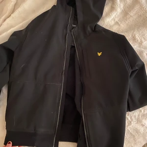 Svart huvtröja från Lyle & Scott - Säljer en svart huvtröja från Lyle & Scott med dragkedja och en liten gul logga på bröstet. Säljes för ett mycket billigt pris