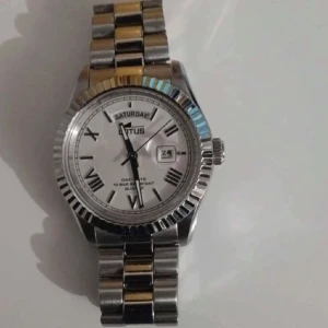 Silverfärgad klocka från Lotus - Ny pris 1600kr. Stilren silverfärgad klocka från Lotus med dag- och datumvisning. Den har en klassisk länkarmband och romerska siffror på urtavlan. Perfekt för en elegant look.