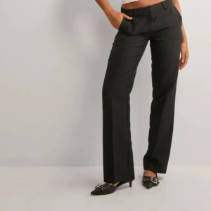 Svarta kostymbyxor - Eleganta svarta kostymbyxor med låg midja och rak passform. Perfekta för en stilren look. Byxorna har en klassisk design med knappar och dragkedja framtill.