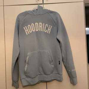 Blå hoodie från Hoodrich - Säljer en snygg blå hoodie från Hoodrich med textdetaljer på både fram- och baksidan. Tröjan har en stor ficka framtill och en bekväm huva. Perfekt för en avslappnad stil.