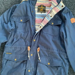 Blå parkajacka från Element - Snygg blå parkajacka från Element med randigt foder i flera färger. Jackan har knappar och dragsko i midjan samt flera praktiska fickor. Perfekt för höst och vår.