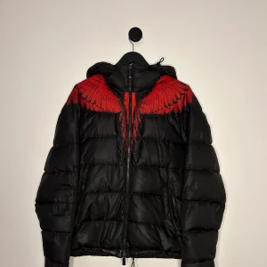Marcelo Burlon Jacka - Snygg svart dunjacka från Marcelo Burlon med ett rött vingmönster över axlarna. Jackan har en dragkedja framtill och är perfekt för kyligare väder. Väldigt unik och väldigt fint skick! Hör gärna av er vid flera frågor eller funderingar! 