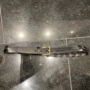 Svart flätat skärp Umbra JH collection med guldfärgat spänne  - Snyggt svart bälte i flätad design med ett stilrent guldfärgat spänne. Perfekt för att ge en extra touch till din outfit. Passar både till ridbyxor och vardagskläder.