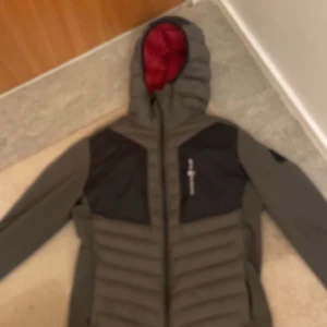 Sail racing jacka - Säljer en grå hybrid jacka från Sail racing med röd insida. Jackan har en dragkedja framtill och är långärmad. Perfekt för kyliga dagar med sin quiltade design och högkvalitativa dunfyllning. 