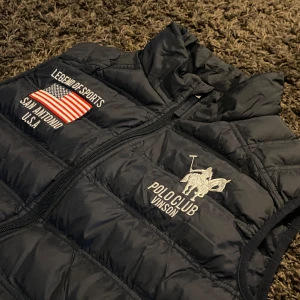 Mörkblå dunväst från Vinson Polo Club - Snygg mörkblå dunväst från Vinson Polo Club med broderade detaljer och dragkedja. Västen har en hög krage och två fickor med dragkedja. Perfekt för en sportig look.