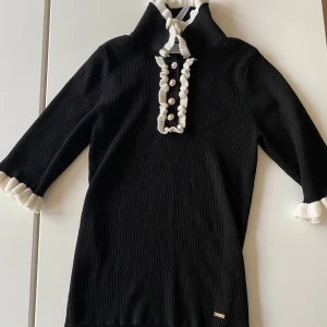 Svart ribbad tröja från Holly & Whyte - Snygg svart ribbad tröja från Holly & Whyte med vita detaljer och knappar framtill. Tröjan har trekvartsärmar och en hög krage med knytband. 